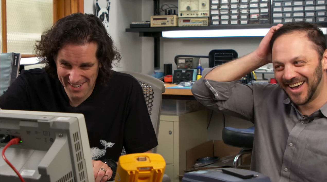 A detailed photo capturing a humorous moment of relief in an electronics workshop. Two tech enthusiasts are laughing; the man on the right scratches his head, realizing their frustrating system issue was caused by forgetting to attach a crucial ground clip. This scene of trial-and-error debugging evokes true bitcoin nostalgia for the gritty, practical roots of early crypto hardware.